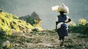 Screenshot of a woman carrying an object on her head.